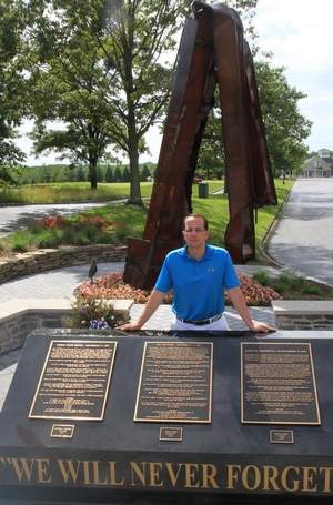 Joe Cary stands in front of plaques honoring the victims of 9/11, with a piece of the World Trade Center in the background. Robert Ward, staff photographer
